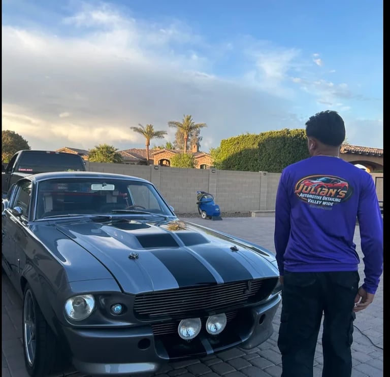Person in purple shirt standing next to a classic black muscle car with white racing stripes in a driveway